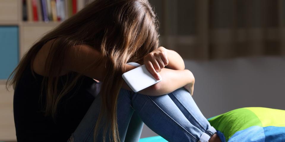 Mädchen hockt auf einem Bett, den Kopf auf die Knie gestützt, in der Hand ein Smartphone.
