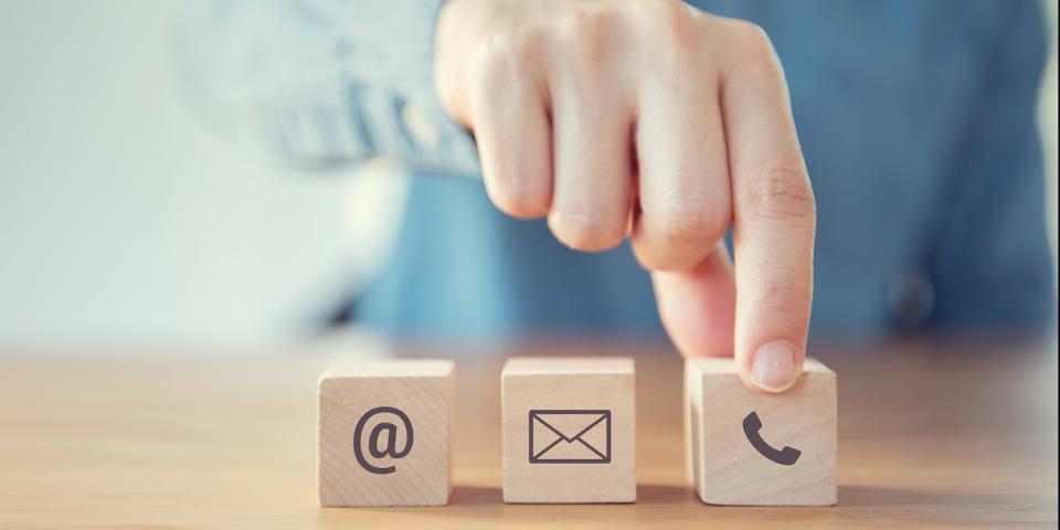 Auf einem Tisch stehen drei hölzerne Würfel nebeneinander, auf denen die Symbole für Telefon, Email und Post gedruckt sind. im Hintergrund der Oberkörper eines Menschen sowie eine ausgestreckte Hand, die nach dem Würfel mit dem Telefon-Symbol greift.