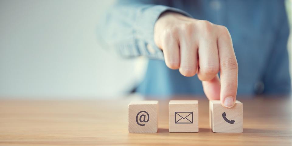 Auf einem Tisch stehen drei hölzerne Würfel nebeneinander, auf denen die Symbole für Telefon, Email und Post gedruckt sind. im Hintergrund der Oberkörper eines Menschen sowie eine ausgestreckte Hand, die nach dem Würfel mit dem Telefon-Symbol greift.