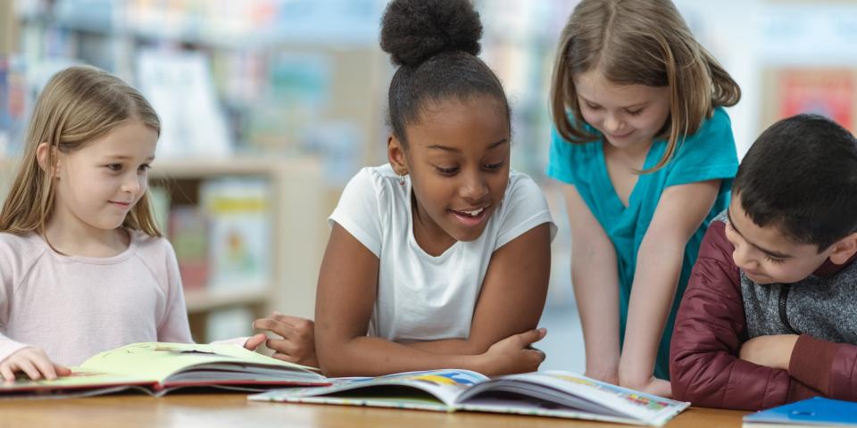 Grundschülerinnen und Grundschüler sitzen an einem Tisch vor aufgeschlagenen Büchern, ein Mädchen liest vor und die anderen hören zu.