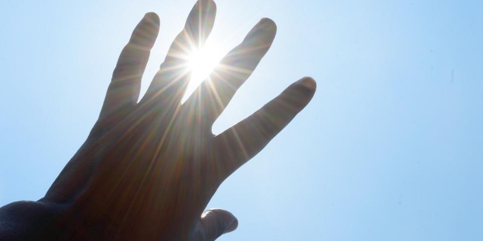 Eine linke Hand vor blauem Himmel, durch die Finger blinzelt die Sonne hindurch.