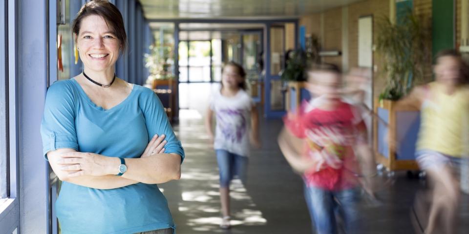 Lehrerin steht lächelnd in einem Schulflur, unscharf  im Hintergrund zu erkennen sind laufende Schülerinnen und Schüler.