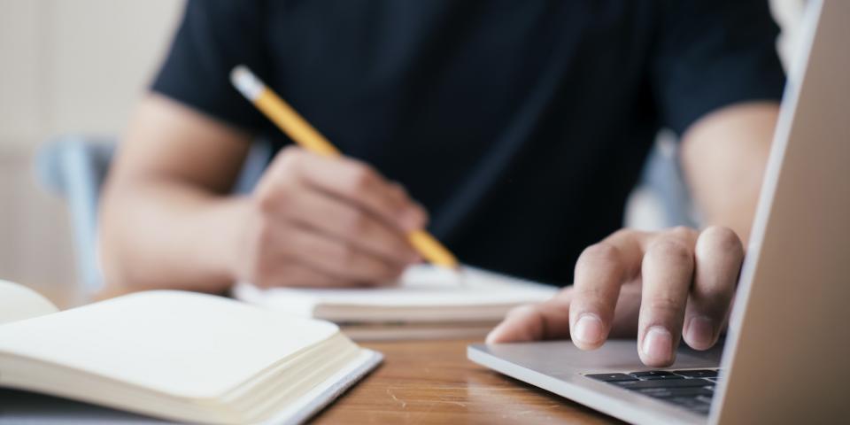 Eine Person, deren Gesicht nicht zu erkennen ist, sitzt an einem Schreibtisch, mit der einen Hand schreibend, die andere Hand liegt auf der Tastatur eines Laptops.