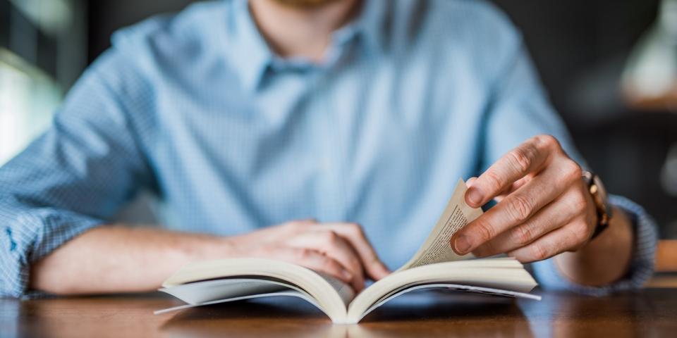 Nahaufnahme: Auf einem Tisch liegt ein geöfffnetes Buch, eine Hand hält es offen, die andere blättert darin. Im Hintergrund ist unscharf die lesende Person zu erkennen.