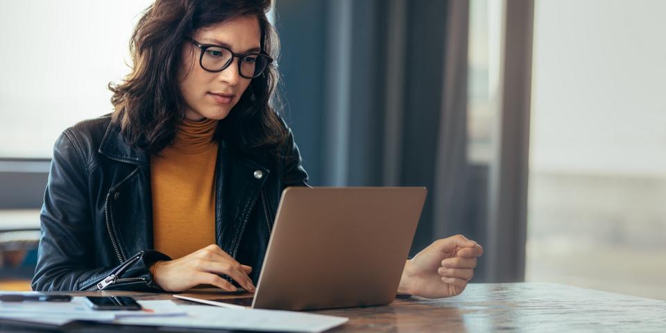 Eine Frau sitzt an einem Schreibtisch und schaut in einen aufgeklappten Laptop.