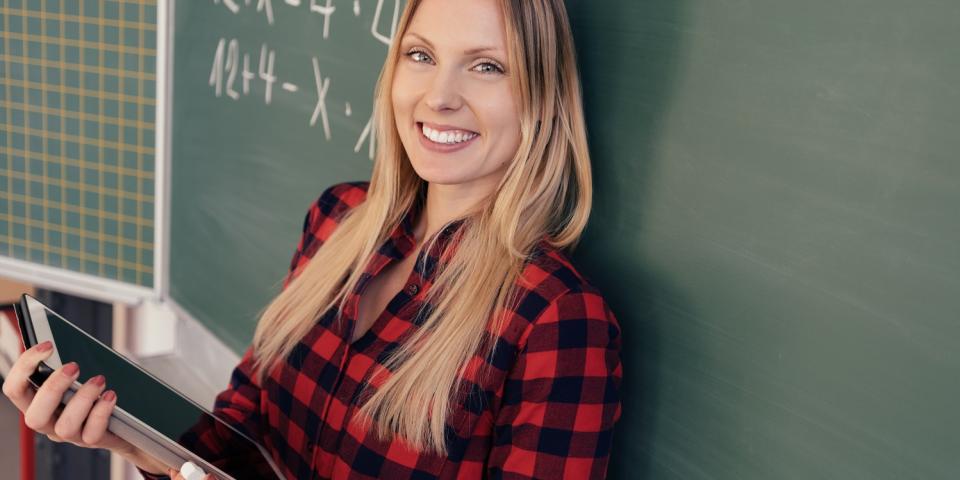 Eine Lehrerin steht mit dem Rücken zur Tafel, in ihren Händen hält sie einen Tablet-Computer.
