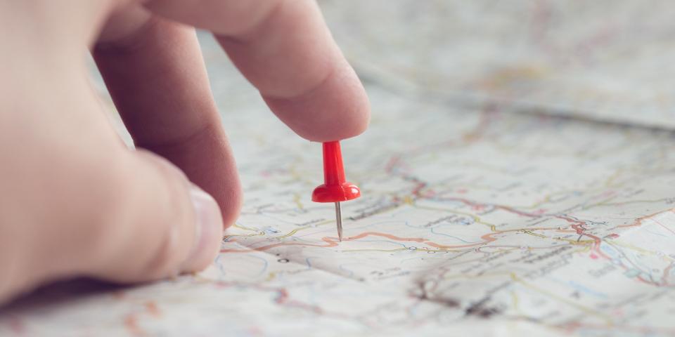 Nahaufnahme einer Landkarte, eine Hand deutet auf einen in die Karte gesteckten Pin.