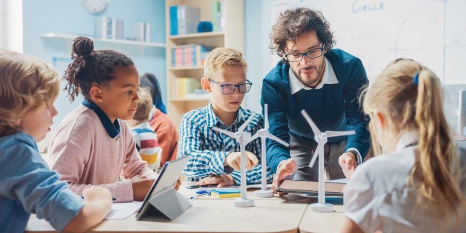 Lehrer mit Schülerinnen und Schülern in einer Unterrichtssituation. Auf dem Tisch stehen Modellwindräder, die die Kinder und der Lehrer gemeinsam betrachten.