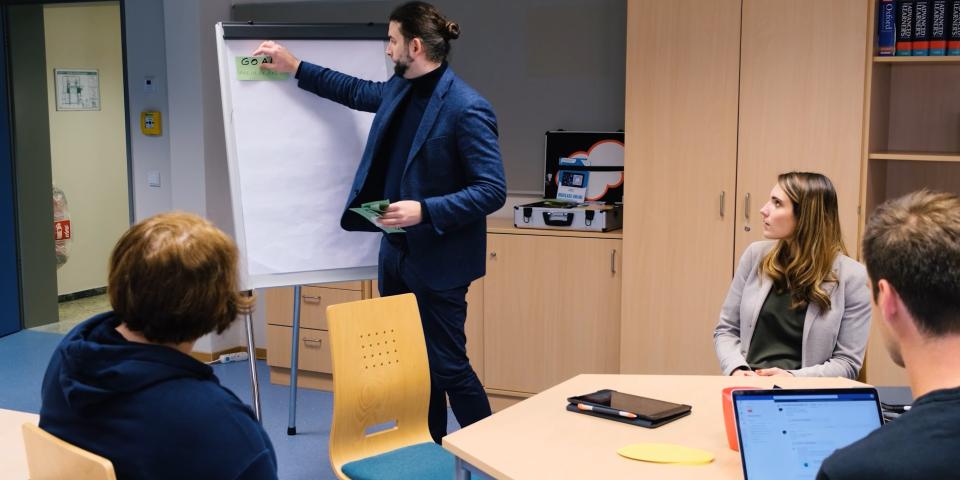 Videostandbild: Medienberatende in einer Gesprächssituation. Ein Medienberater heftet Moderationskarten an eine Flipchart.