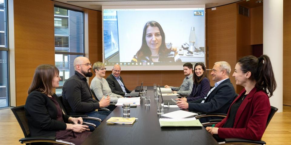 Personen sitzen in einem Besprechungsraum um einen Tisch, wobei über eine Leinwand eine externe Besprechungsteilnehmerin eingeblendet wird