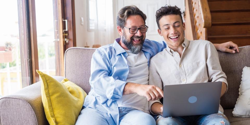 Vater und Sohn sitzen auf einem Sofa und schauen lächelnd in einen geöffneten Laptop auf dem Schoß des Sohnes.