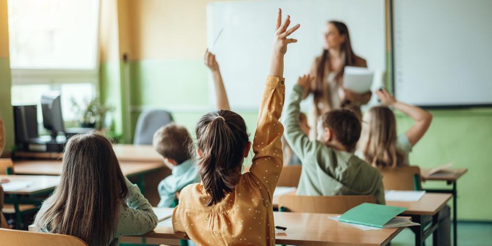 Rückansicht von Grundschülerinnen und Grundschülern, die in einem Klassenraum an ihren Tischen sitzen. Im Hintergrund ist unscharf eine Lehrerin zu erkennen.