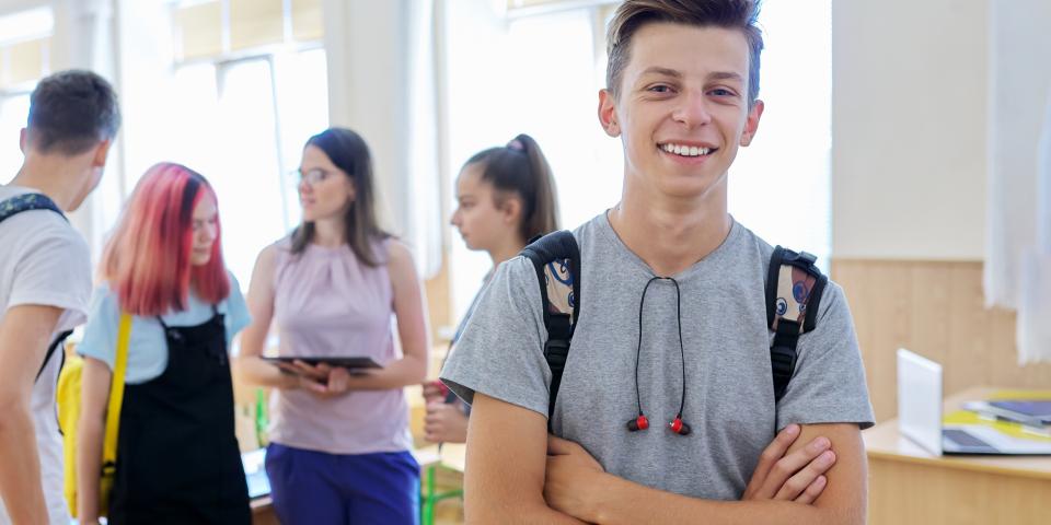 Eine Gruppe Schülerinnen und Schüler steht in einem Klassenraum und spricht miteinander. Ein Jugendlicher im Vordergrund blickt lächelnd in die Kamera.