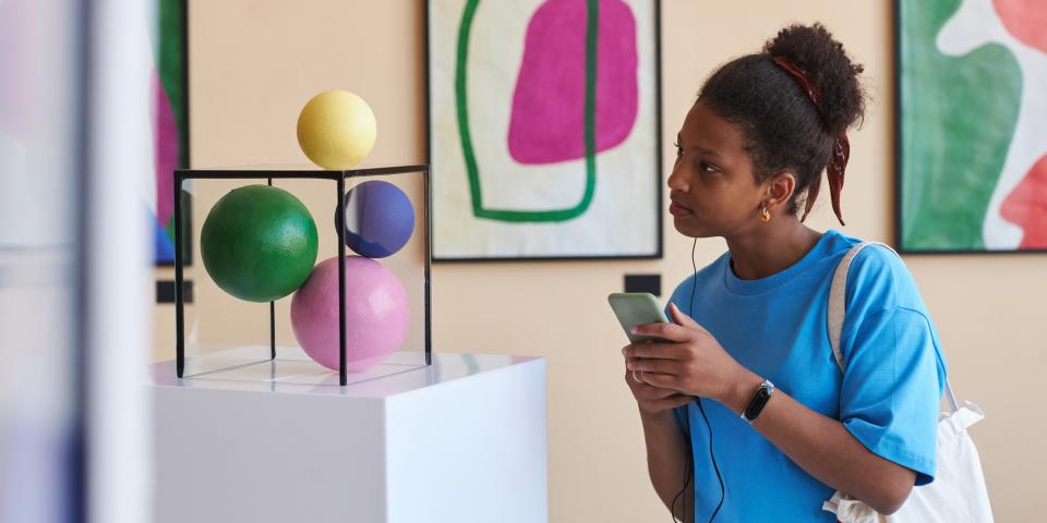 Eine Schülerin steht in einem Museum mit dem Handy in der Hand und einem Kopfhörer im Ohr vor einer Skulptur.