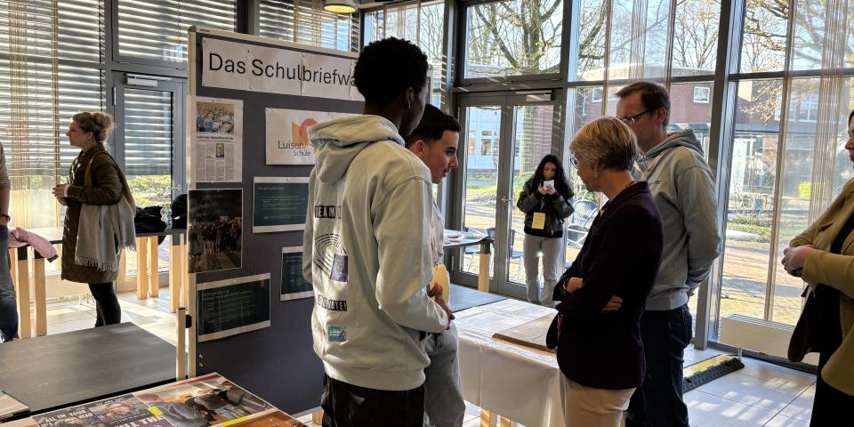 Schulministerin Feller steht mit Schülerinnen und Schülern vor einem Stand der Regionalen Lernstatt NRW über die Schulbriefwahl.