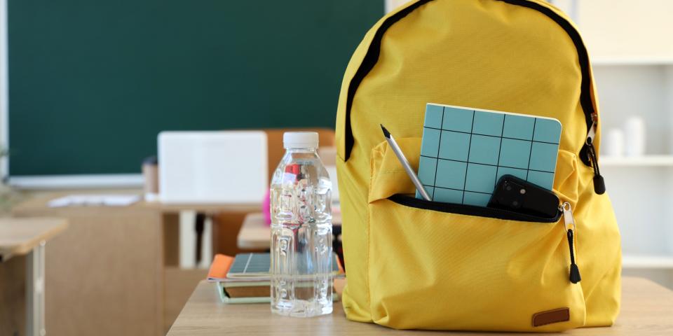 Symbolbild: Rucksack steht auf einem Schultisch, ein Smartphone und Schreibutensilien ragen aus einer geöffneten Tasche.