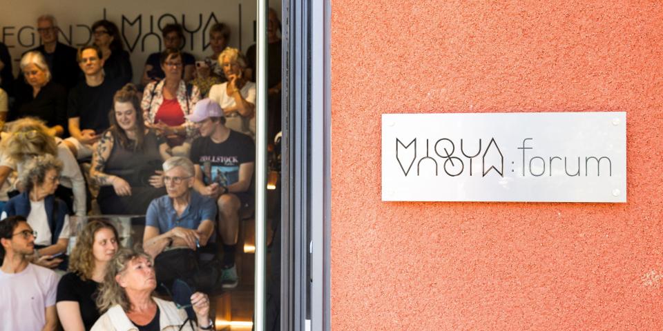 Durch die geöffnete Tür blickt man ins MiQua.forum, in dem vielen Menschen an einer Veranstaltung teilnehmen.