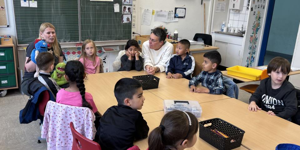 Lehrerin sitzt mit einer Handpuppe vor Kindern, die an einem Tisch sitzen.