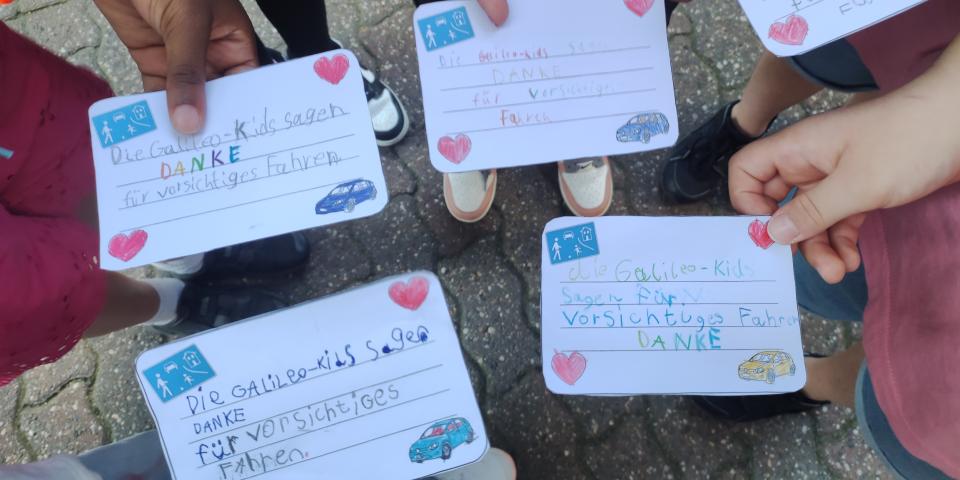 Mehrere Kinderhände halten beschriftete Karten zum Thema Straßenverkehr in die Bildmitte.