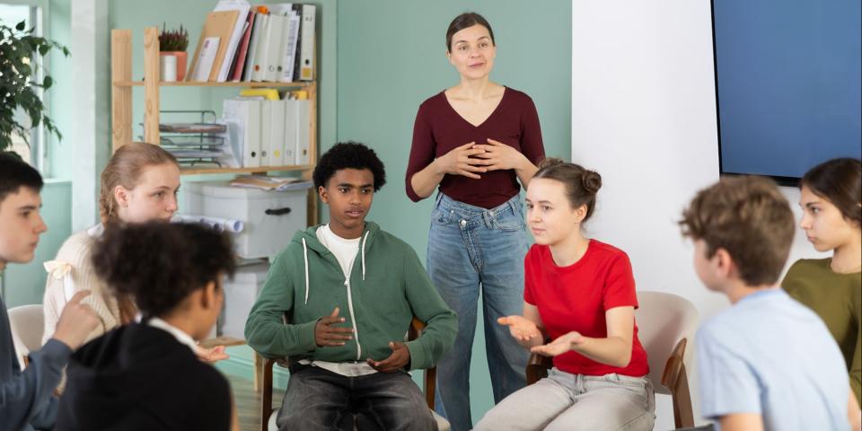 Schülerinnen und Schüler sitzen im Kreis und diskutieren, eine Lehrerin beobachtet die Diskussion im Hintergrund.