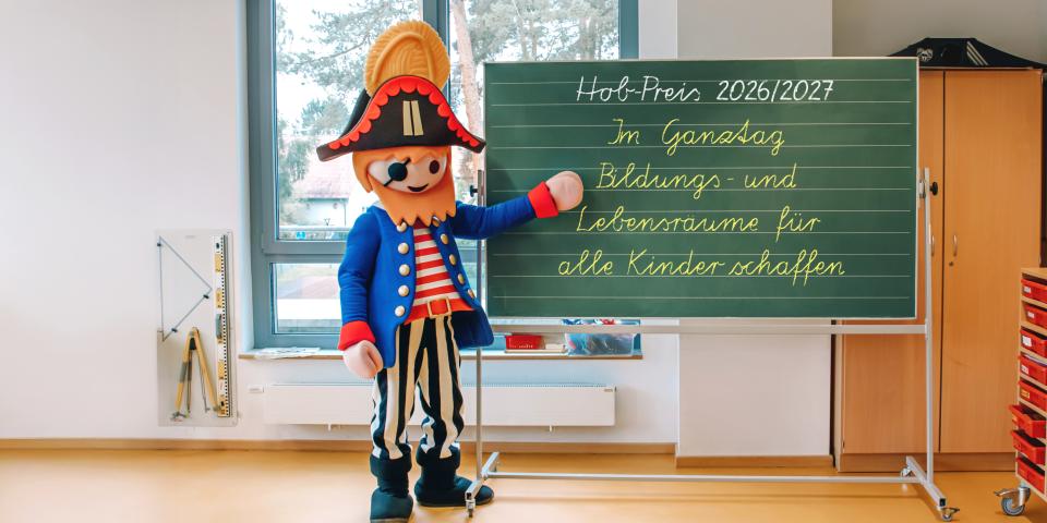 Ein Playmobil-Pirat steht an einer Tafel mit der Aufschrift "Hob-Preis 2026/27. Im Ganztag Bildungs- und Lebensräume für alle Kinder schaffen".