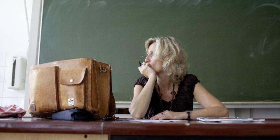 Lehrerin sitzt nachdenklich am Tisch vor einer Tafel.