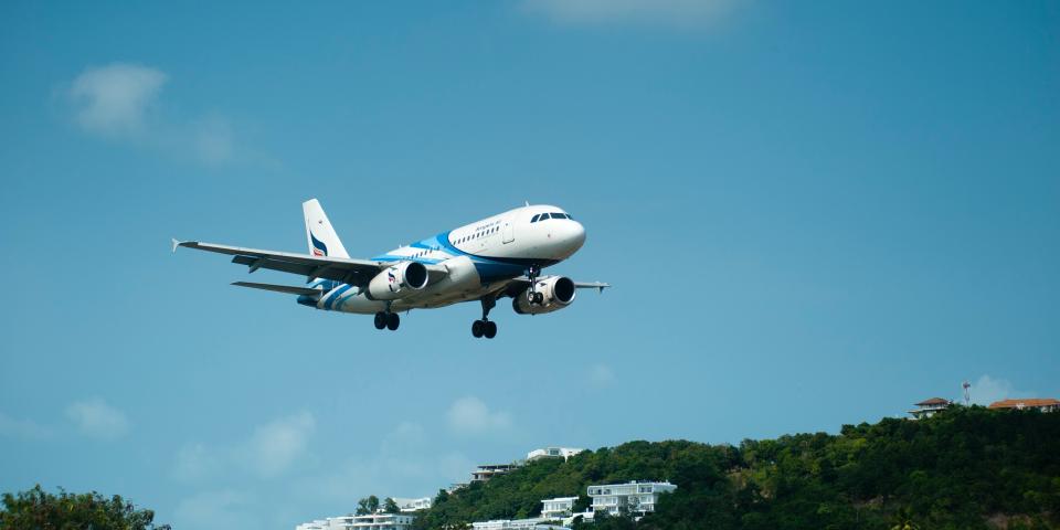 Blauer Himmel, davor startendes oder landendes Flugzeug, im Hintergrund bewaldete Landschaft mit weißen Häusern.