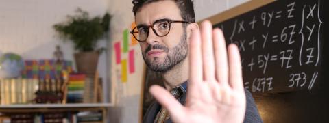 Lehrer steht mit zur Abwehr erhobener Hand vor der Tafel.