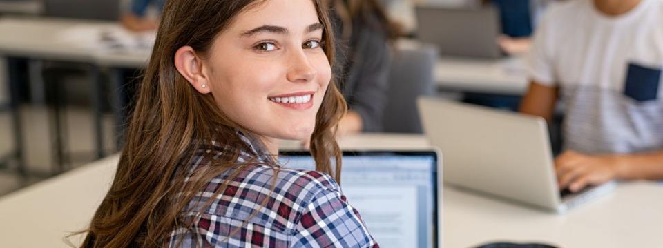 Eine Schülerin sitzt in einer Unterrichtssituation am Laptop und wendet sich dem Betrachter zu.