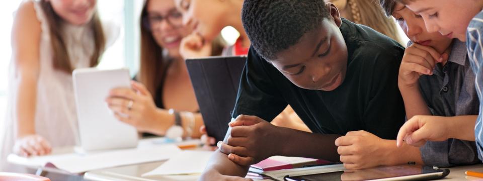 Schülerinnen und Schüler blicken auf Tablets, die vor ihnen auf dem Tisch liegen.