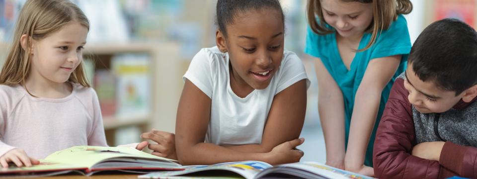 Grundschülerinnen und Grundschüler sitzen an einem Tisch vor aufgeschlagenen Büchern, ein Mädchen liest vor und die anderen hören zu.