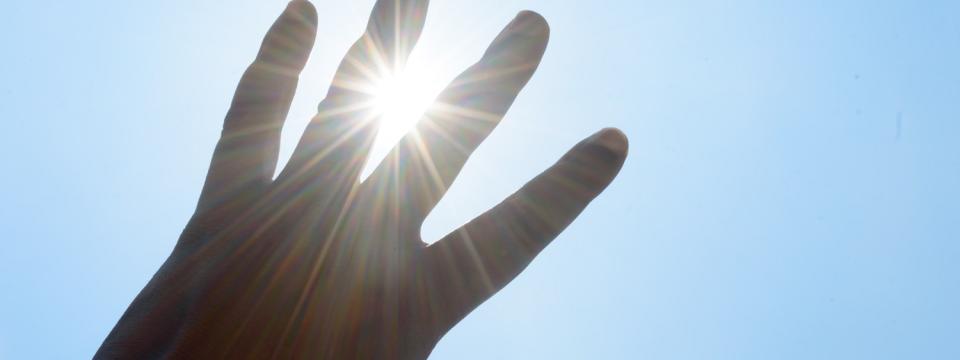Eine linke Hand vor blauem Himmel, durch die Finger blinzelt die Sonne hindurch.