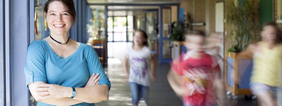 Lehrerin steht lächelnd in einem Schulflur, unscharf  im Hintergrund zu erkennen sind laufende Schülerinnen und Schüler.