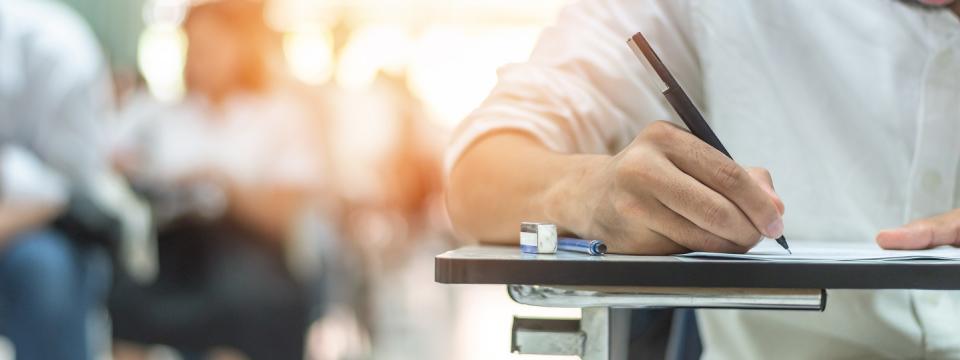 Detailaufnahme einer Hand, die auf Papier schreibt. Im Hintergrund unscharf weitere schreibende Personen an Tischen.