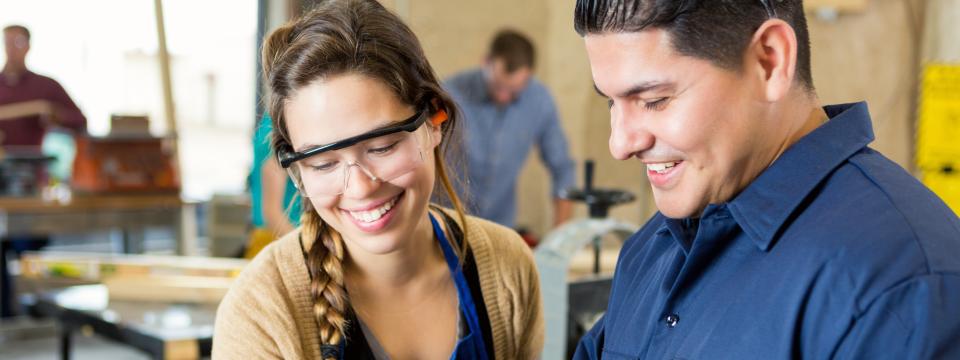 Eine Schülerin und ein Lehrer stehen zusammen in einer Werkstatt, sie tragen Arbeitskleidung und Schutzbrillen.