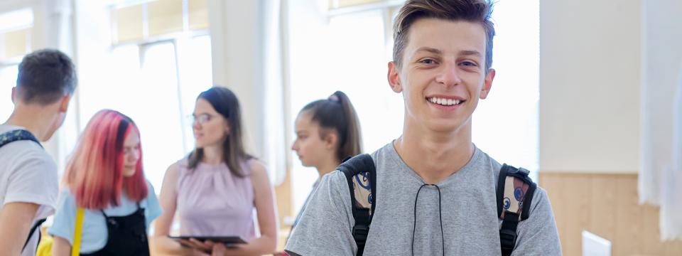 Eine Gruppe Schülerinnen und Schüler steht in einem Klassenraum und spricht miteinander. Ein Jugendlicher im Vordergrund blickt lächelnd in die Kamera.
