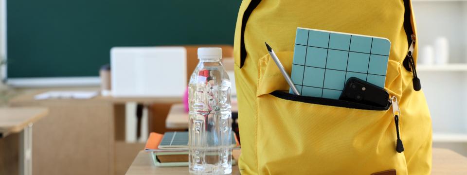 Symbolbild: Rucksack steht auf einem Schultisch, ein Smartphone und Schreibutensilien ragen aus einer geöffneten Tasche.