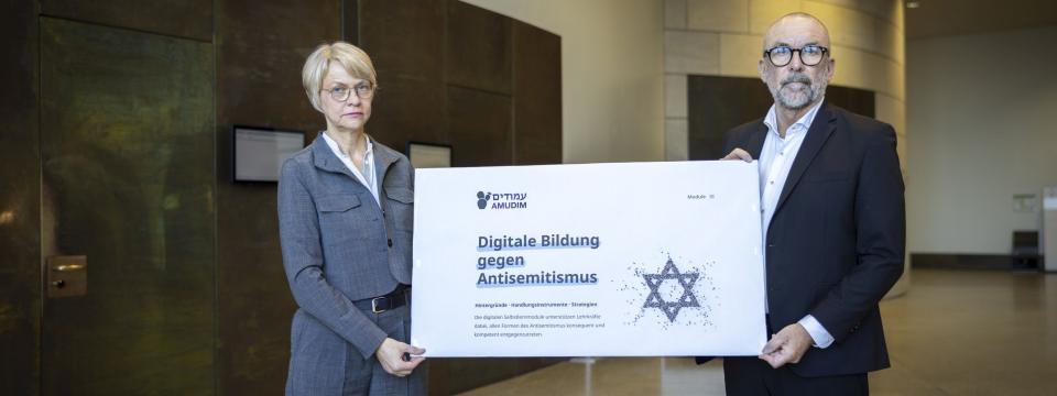 Ministerin Feller und Bert Römgens präsentieren ein Plakat zum Start von AMUDIM (25. Februar 2026)