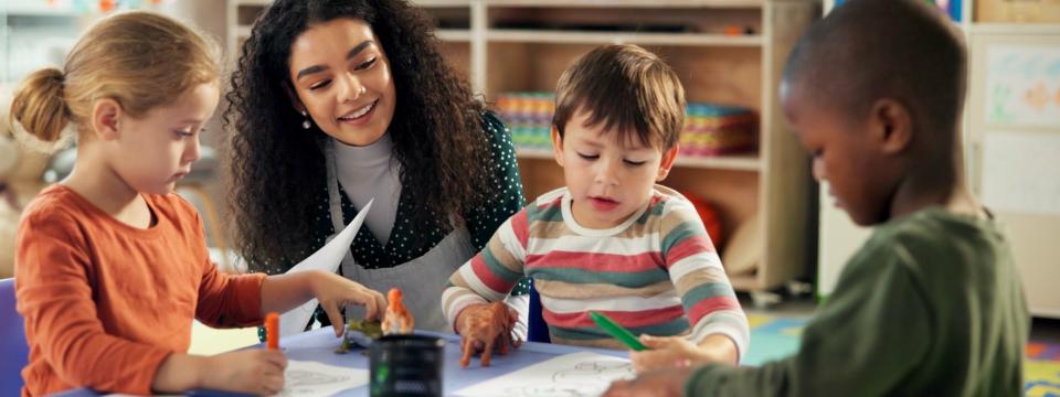 Symbolbild: Lehrerin spricht mit einer Gruppe Kinder, die an einem Tisch sitzen.