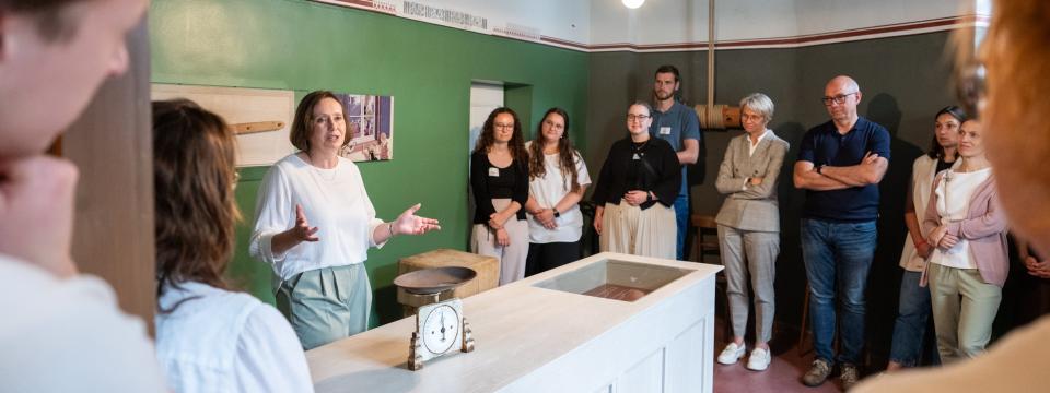 Ministerin Feller sowie Lehramtsanwärterinnen und -anwärter stehen in einem Raum des Humberghauses und hören einer Führung zu.