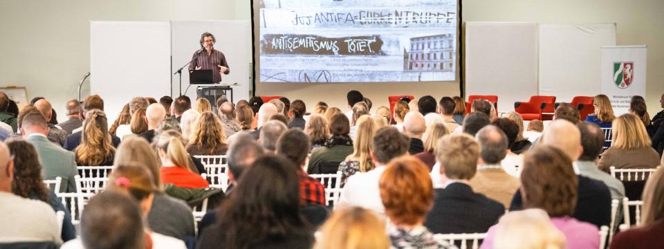 Ein Teilnehmer steht an einem Rednerpult, neben ihm eine Beamerpräsentation, im Saal sitzen zahlreiche Zuhörerinnen und Zuhörer.