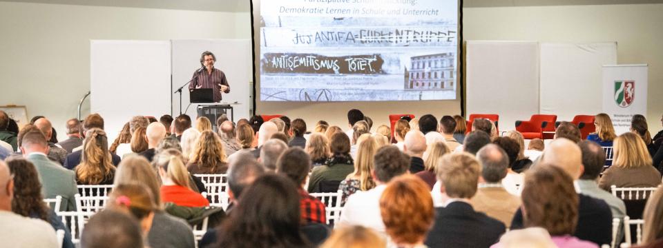 Ein Teilnehmer steht an einem Rednerpult, neben ihm eine Beamerpräsentation, im Saal sitzen zahlreiche Zuhörerinnen und Zuhörer.