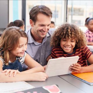 Lehrer, eine Schülerin und ein Schüler blicken in einem Klassenraum gemeinsam auf ein Tablet (Computer). 