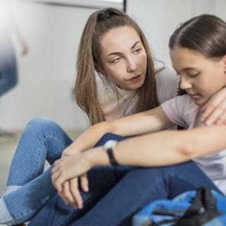 Eine junge Frau tröstet ein Mädchen, beide sitzen auf dem Boden, die eine hat den Arm um die andere gelegt.