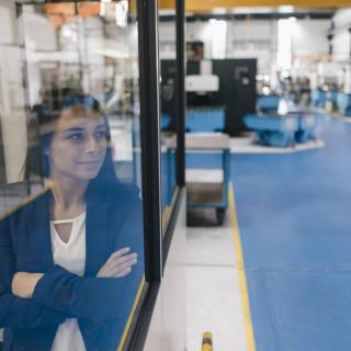 Eine Frau blickt durch eine Glasscheibe in eine High-Tech-Produktionshalle.