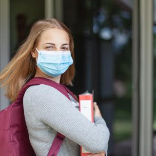 Eine Schülerin mit Maske trägt ihren Schulrucksack auf dem Rücken und einen Ordner in der Hand und blickt in die Kamera