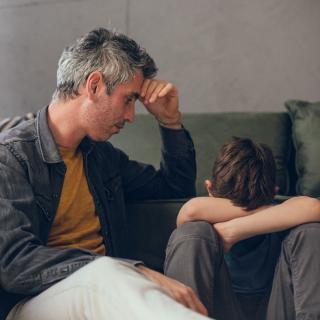 Vater tröstet seinen Sohn der mit dem Kopf auf den Knien neben ihm hockt.