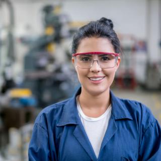 Junge Frau in Arbeitskleidung und Schutzbrille, im Hintergrund Maschinen.