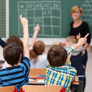 Grundschülerinnen und -schüler melden sich, im Hintergrund eine Tafel mit Rechenaufgaben