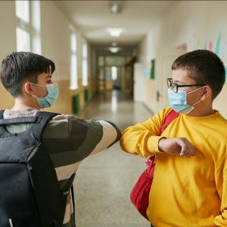 Zwei Alltagsmasken tragende Schüler begrüßen sich mit einem Ellenbogencheck auf einem Schulflur.